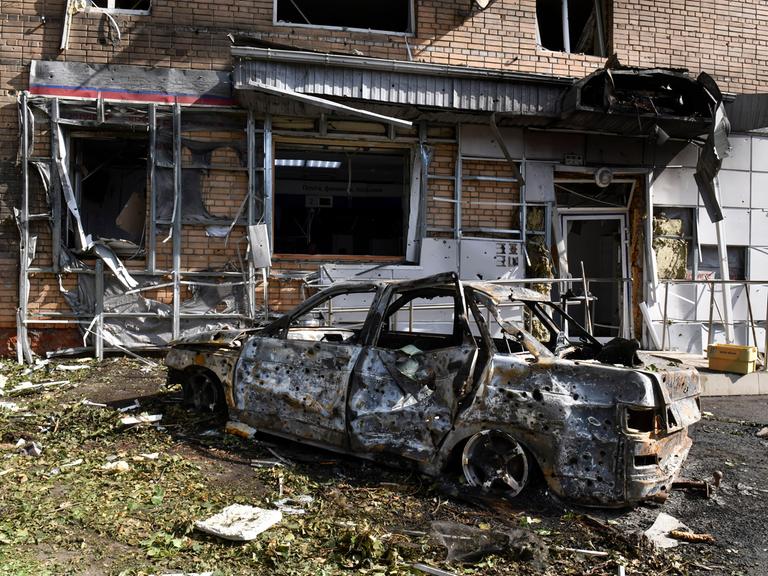 Ein ausgebranntes Auto steht vor einem nach Angriffen zerstörten Gebäude