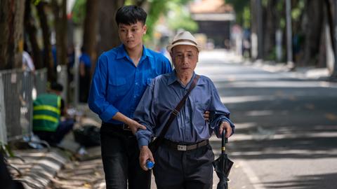 Ein junger Vietnamese hält einen älteren Mann am Arm und hilft ihm auf der Straße. (Symbolbild)