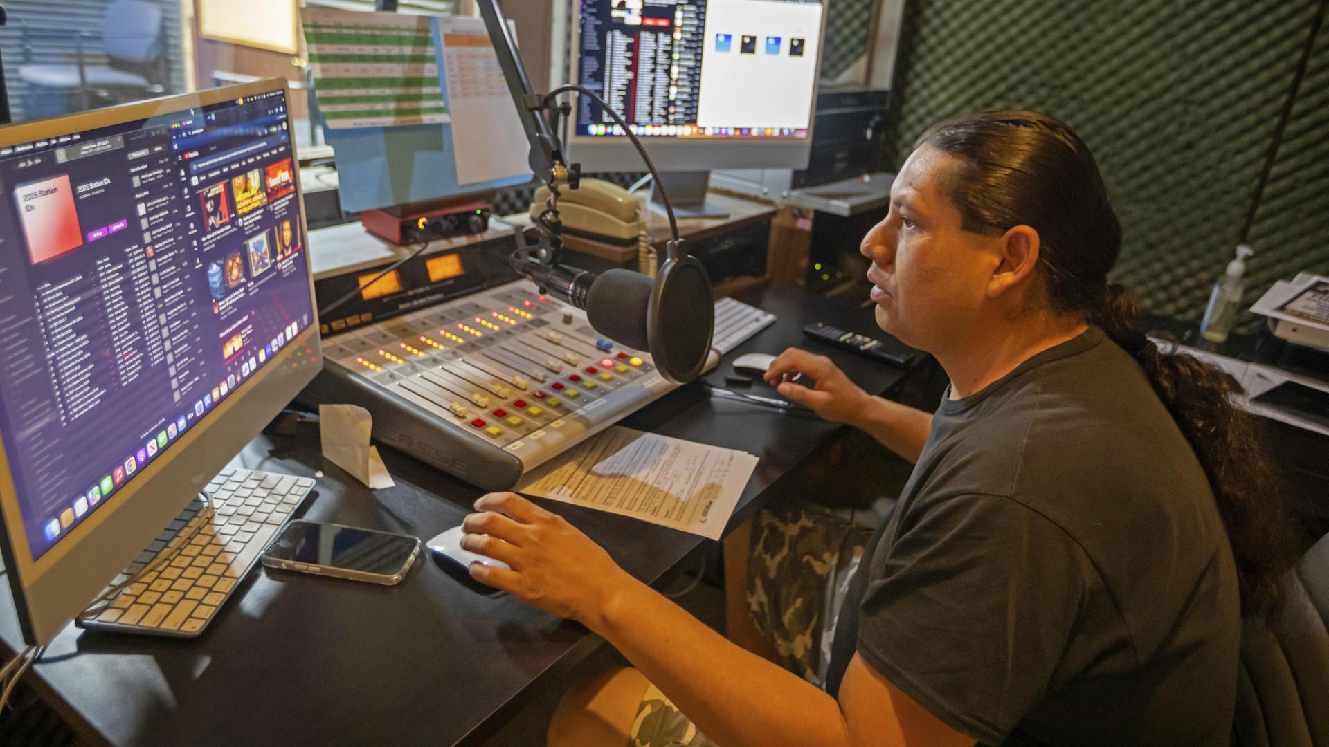 Der Moderator mit dunklem Pferdeschwanz und dunklem T-Shirt sitzt in einem kleinen Selbsfahrerstudio vor dem Mikrofon und bedient per Maus einen Bildschirm.
