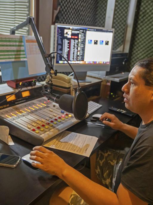 Der Moderator mit dunklem Pferdeschwanz und dunklem T-Shirt sitzt in einem kleinen Selbsfahrerstudio vor dem Mikrofon und bedient per Maus einen Bildschirm.