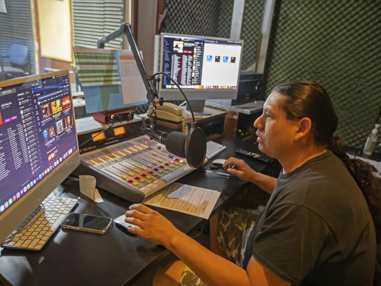 Der Moderator mit dunklem Pferdeschwanz und dunklem T-Shirt sitzt in einem kleinen Selbsfahrerstudio vor dem Mikrofon und bedient per Maus einen Bildschirm.