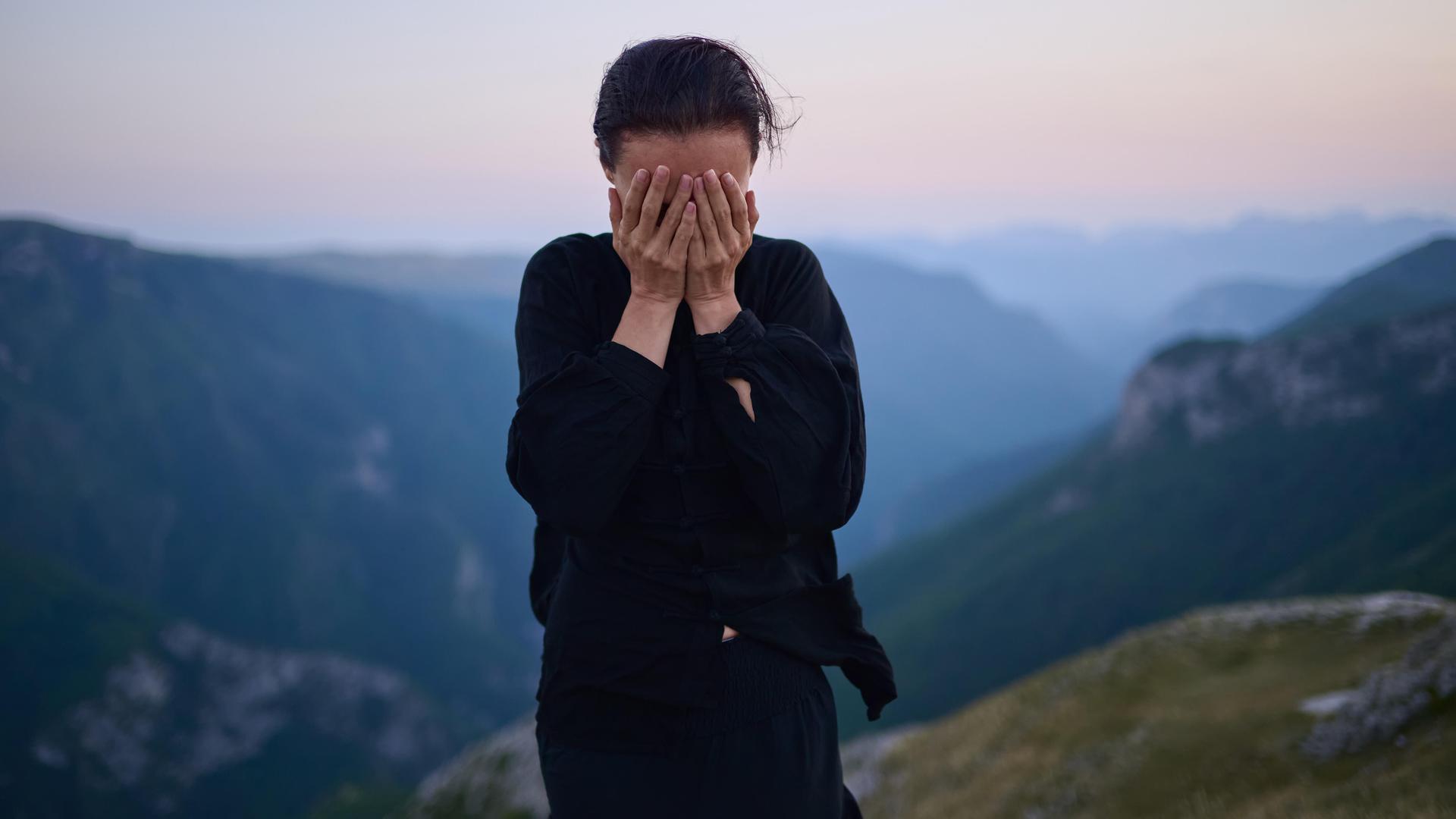 Eine Frau steht in traditioneller schwarzer Tai-Chi-Kleidung auf einem Berg bei Sonnenaufgang. Sie hat die Hände vor das Gesicht geschlagen.