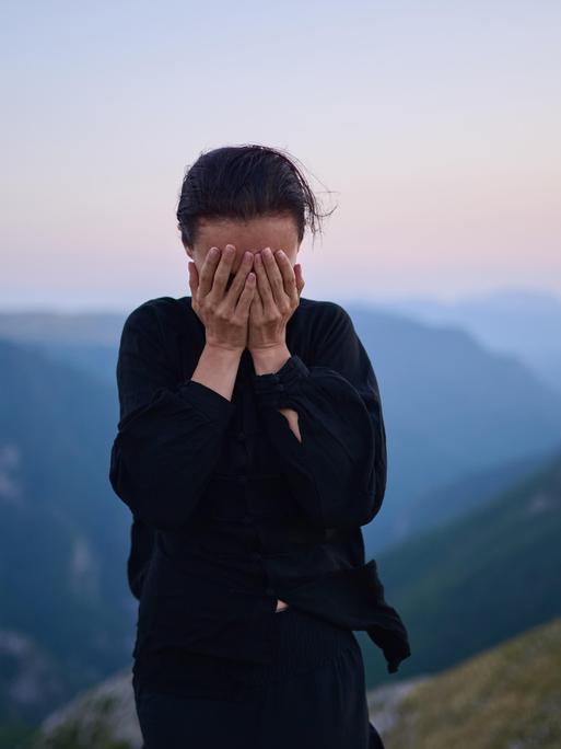 Eine Frau steht in traditioneller schwarzer Tai-Chi-Kleidung auf einem Berg bei Sonnenaufgang. Sie hat die Hände vor das Gesicht geschlagen.