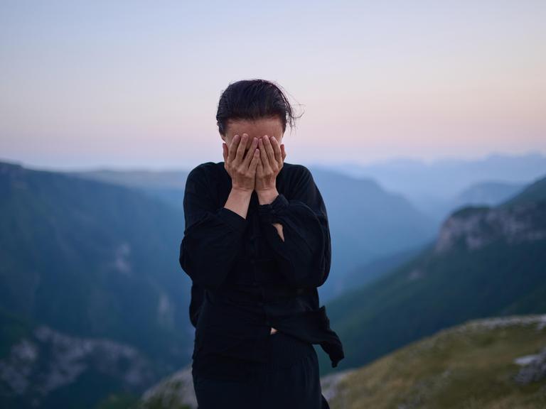 Eine Frau steht in traditioneller schwarzer Tai-Chi-Kleidung auf einem Berg bei Sonnenaufgang. Sie hat die Hände vor das Gesicht geschlagen.