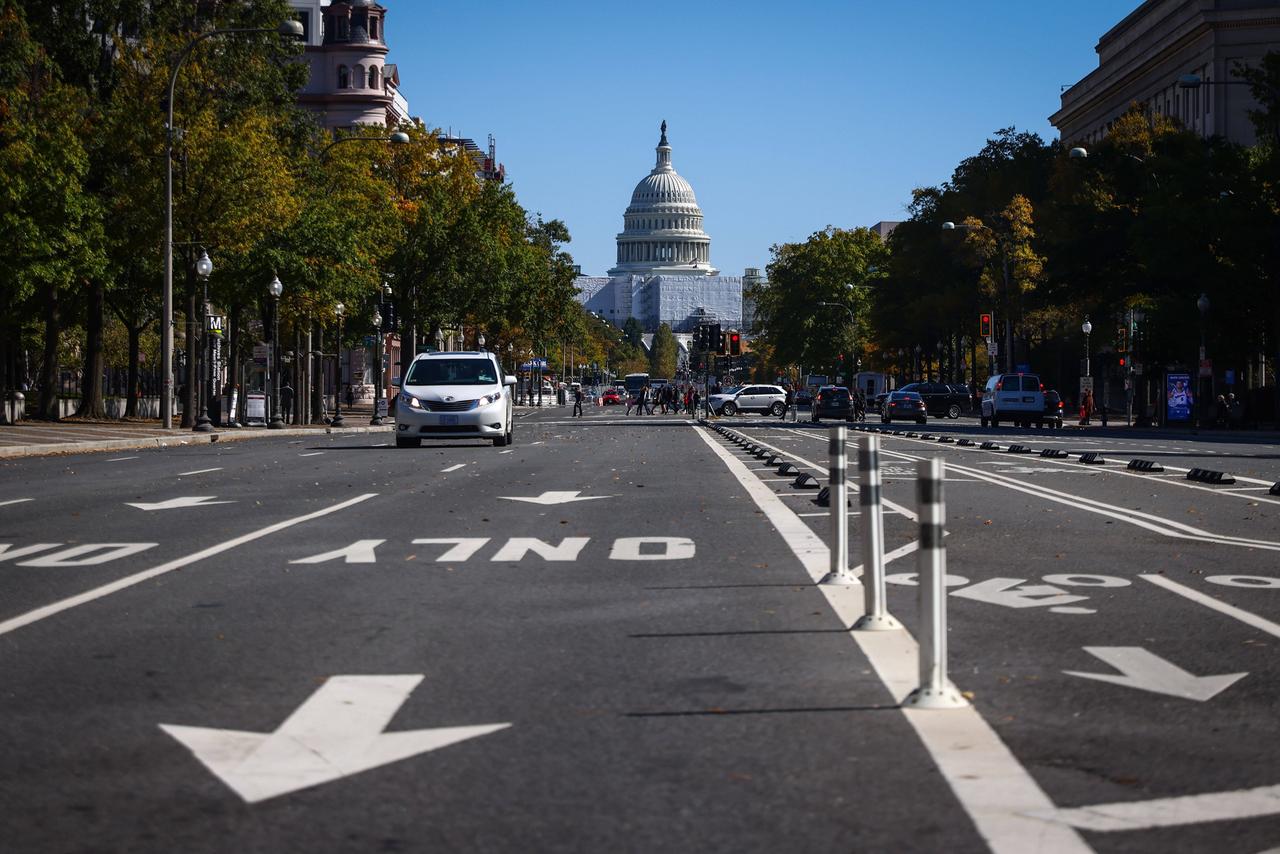 Das Capitol in Washington D.C. - im Vordergrund eine Straße mit Autos, im Hintergrund das Gebäude vor blauem Himmel. Die Straße wird von Bäumen gesäumt.