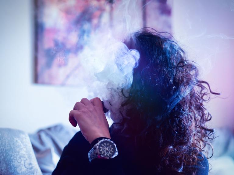 Eine junge Frau raucht eine E-Zigarette. Ihr Gesicht verschwindet hinter dem Dampf.