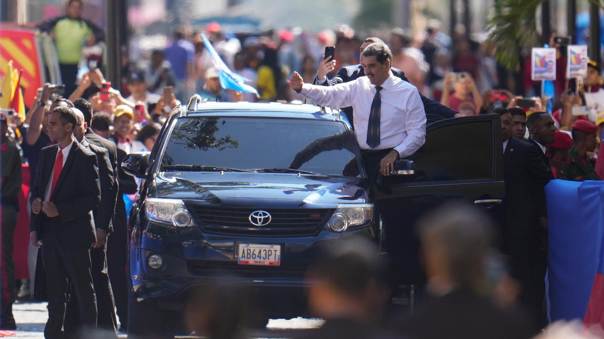 Blick aus größerer Entfernung: Maduro im weißen Hemd und mit blauer Krawatte steigt aus einem schwarzen Wagen und winkt in die jubelnde Menge drumherum.