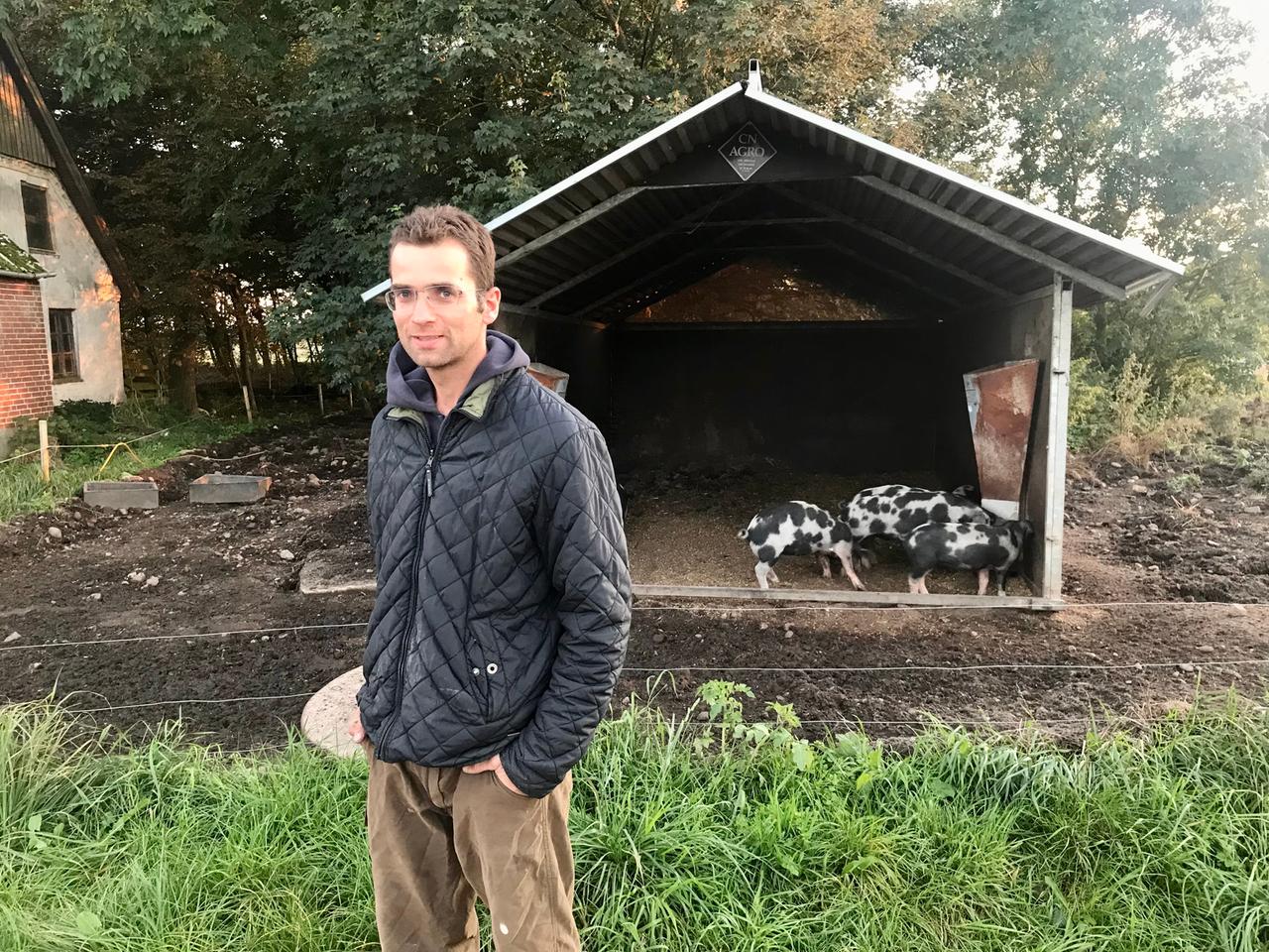 Ein Mann in dunklem Anorak steht vor einem Stall mit  scheckigen Schweinen.