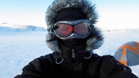 Mann mit Kapuze und Gesichtsschutz auf Gletscher in Island, im Hintergrund ein Zelt. Vom Gesicht ist nur die rote Schneebrille zu sehen. 