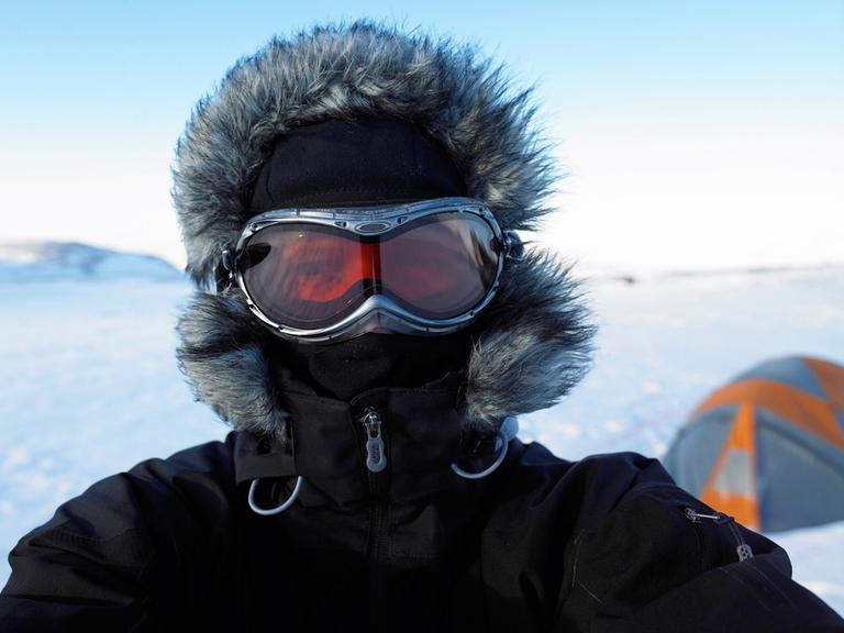 Mann mit Kapuze und Gesichtsschutz auf Gletscher in Island, im Hintergrund ein Zelt. Vom Gesicht ist nur die rote Schneebrille zu sehen. 