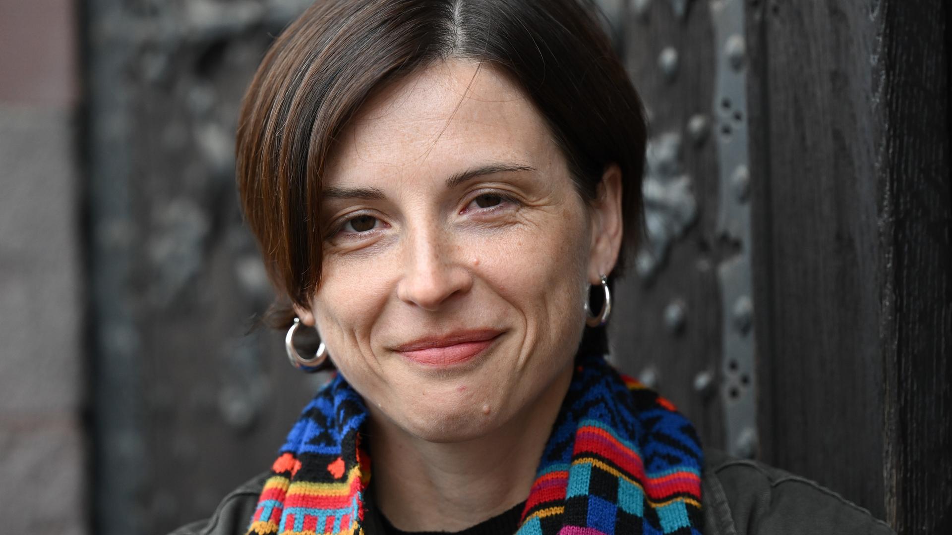 Dorothee Elmiger steht vor einer gusseisernen Tür, trägt einen bunten Schal und lächelt in die Kamera