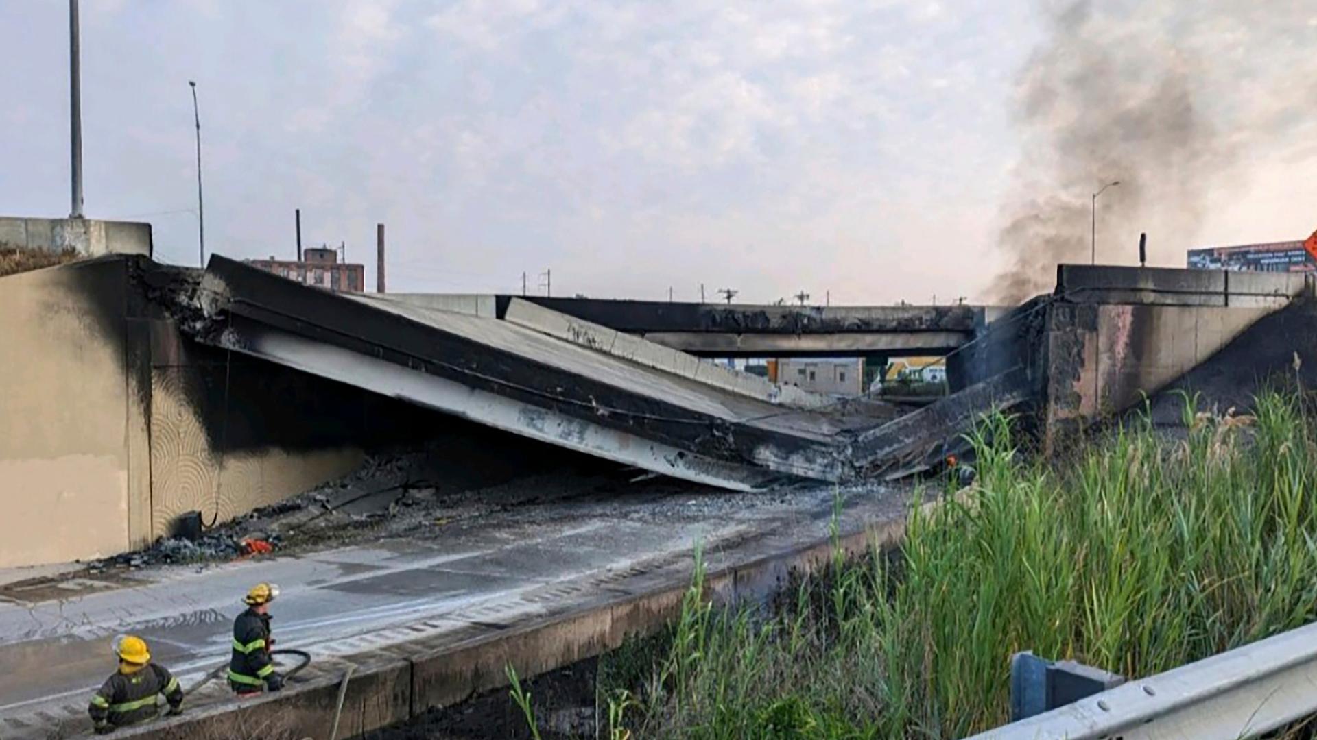 Feuerwehrleute stehen neben einer eingestürzten Autobahnbrücke. Der Beton ist schwarz verfärbt, Rauch steigt auf.