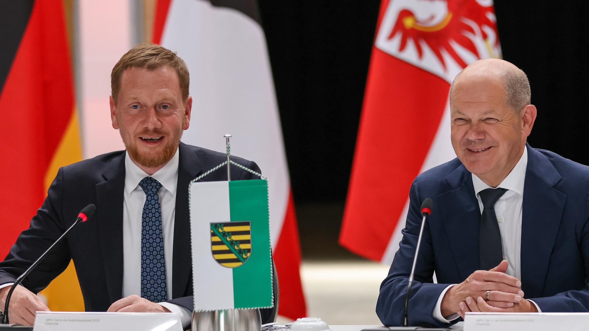 Michael Kretschmer (l, CDU), Ministerpräsident von Sachsen, und Bundeskanzler Olaf Scholz (SPD) sitzen bei der Tagung der Ost-Ministerpräsidenten. Sie lächeln in die Kamera.