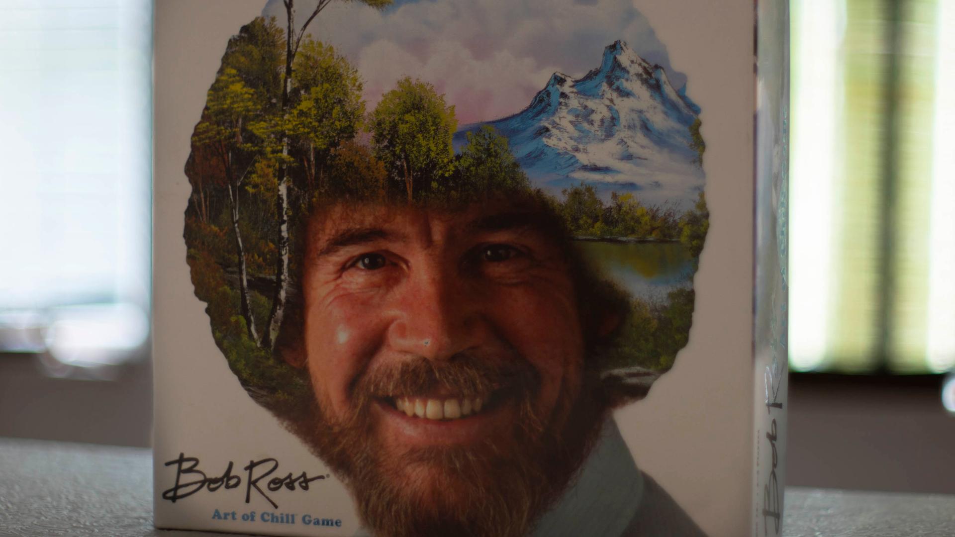 Das Bob Ross-Brettspiel „The Art of Chill“ steht aufrecht auf einem Tisch. Auf dem Deckel ist das Konterfei von Bob Ross zu sehen. Sein lockiges Haar ist mit einer Landschaft mit Bäumen, Bergen, einem See und weißen Wolken am blauen Himmel übermalt.