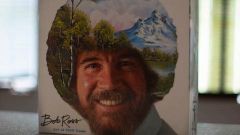 Das Bob Ross-Brettspiel „The Art of Chill“ steht aufrecht auf einem Tisch. Auf dem Deckel ist das Konterfei von Bob Ross zu sehen. Sein lockiges Haar ist mit einer Landschaft mit Bäumen, Bergen, einem See und weißen Wolken am blauen Himmel übermalt.
