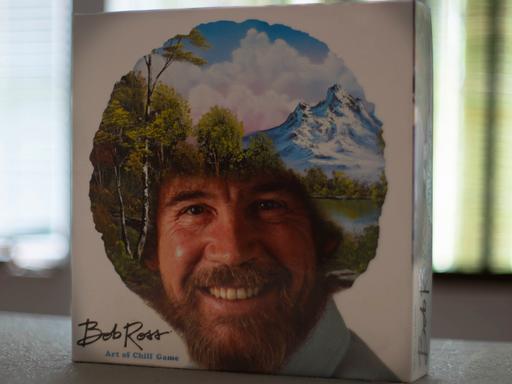 Das Bob Ross-Brettspiel „The Art of Chill“ steht aufrecht auf einem Tisch. Auf dem Deckel ist das Konterfei von Bob Ross zu sehen. Sein lockiges Haar ist mit einer Landschaft mit Bäumen, Bergen, einem See und weißen Wolken am blauen Himmel übermalt.