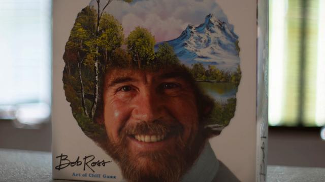 Das Bob Ross-Brettspiel „The Art of Chill“ steht aufrecht auf einem Tisch. Auf dem Deckel ist das Konterfei von Bob Ross zu sehen. Sein lockiges Haar ist mit einer Landschaft mit Bäumen, Bergen, einem See und weißen Wolken am blauen Himmel übermalt.