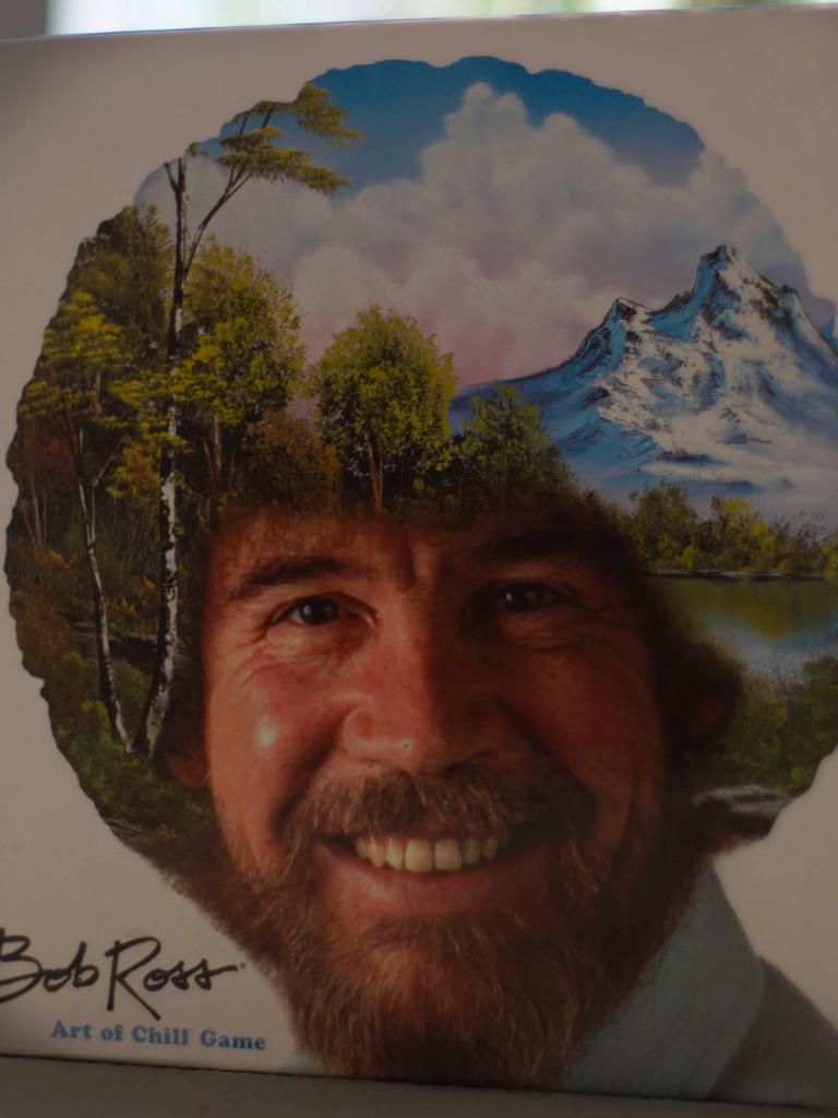 Das Bob Ross-Brettspiel „The Art of Chill“ steht aufrecht auf einem Tisch. Auf dem Deckel ist das Konterfei von Bob Ross zu sehen. Sein lockiges Haar ist mit einer Landschaft mit Bäumen, Bergen, einem See und weißen Wolken am blauen Himmel übermalt.
