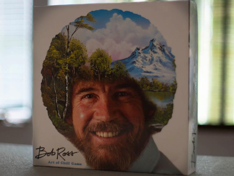 Das Bob Ross-Brettspiel „The Art of Chill“ steht aufrecht auf einem Tisch. Auf dem Deckel ist das Konterfei von Bob Ross zu sehen. Sein lockiges Haar ist mit einer Landschaft mit Bäumen, Bergen, einem See und weißen Wolken am blauen Himmel übermalt.