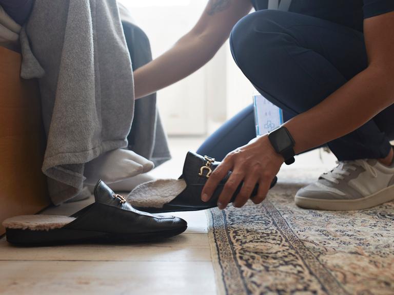 Eine jüngere Person hilft einer älteren Person beim Schuhe anziehen.