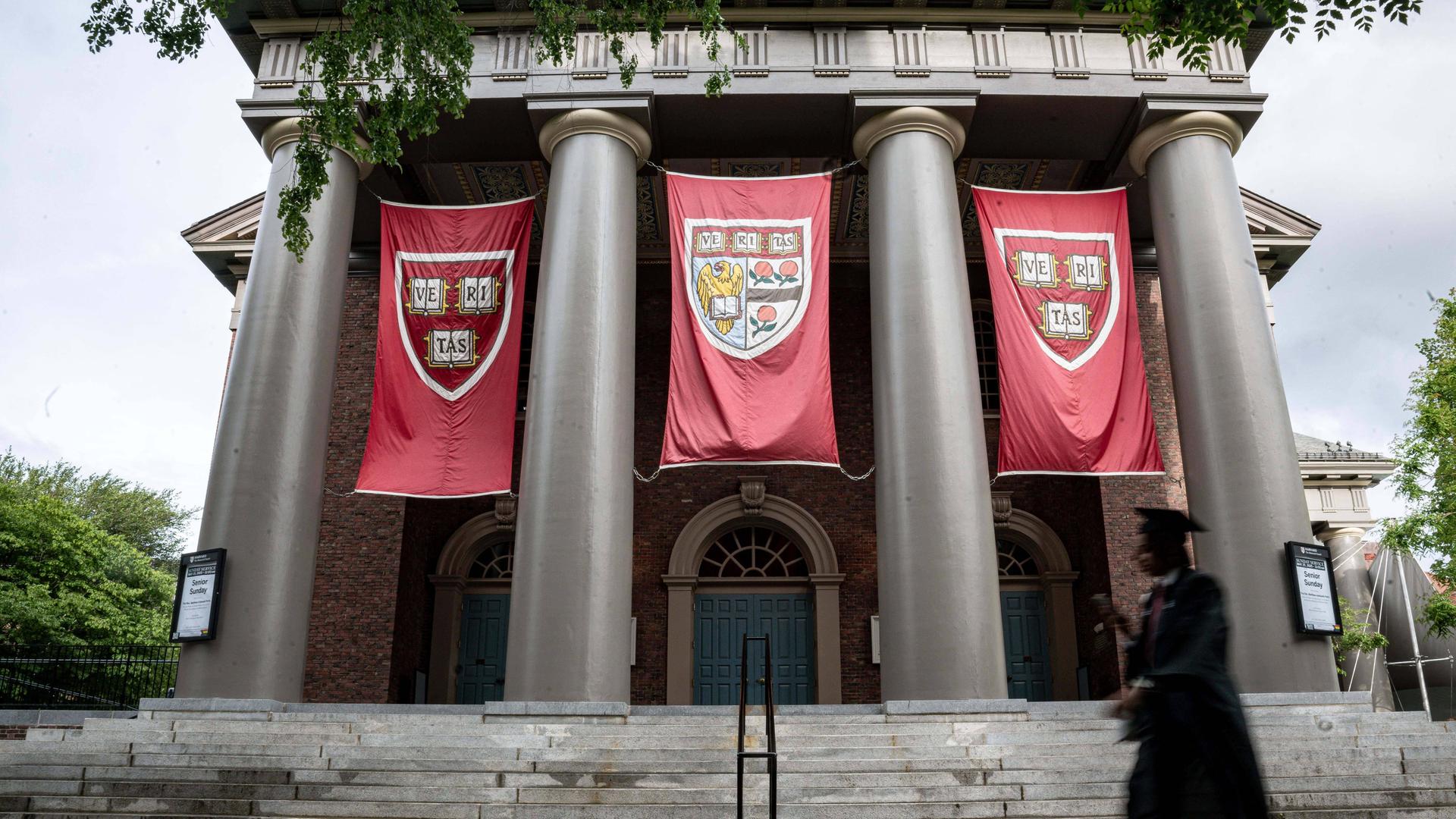 Das Foto zeigt die Appleton Kapelle auf dem Campus der Harvard University in Cambridge, Massachusetts, in den Vereinigten Staaten, mit ihren prächtigen Säulen. Im Vordergrund ist verschwommen die Silhouette einer männlichen Person mit Doktorhut.