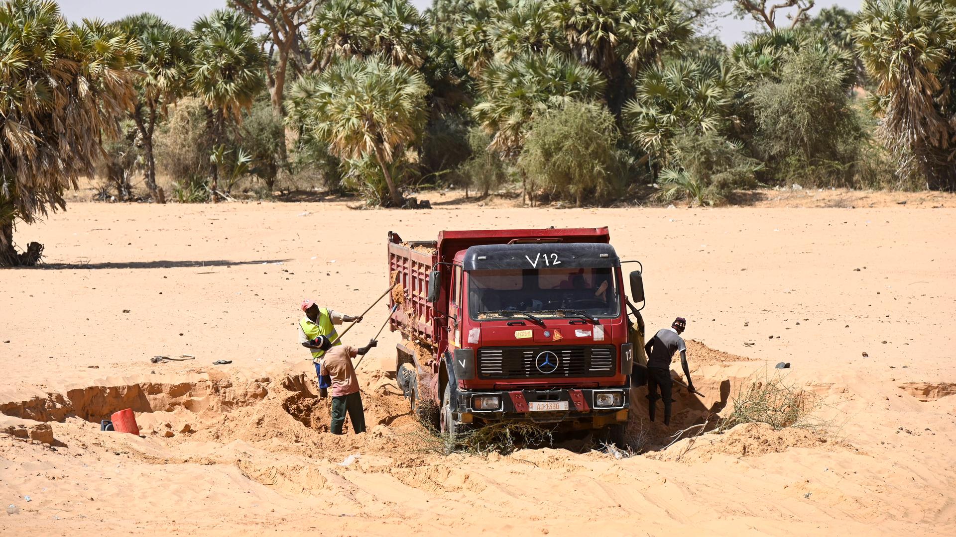 Ein Lastwagen auf sandigem Untergrund, Arbeiter schaufeln Sand in das Fahrzeug. Im Hintergrund sind Bäume zu sehen.