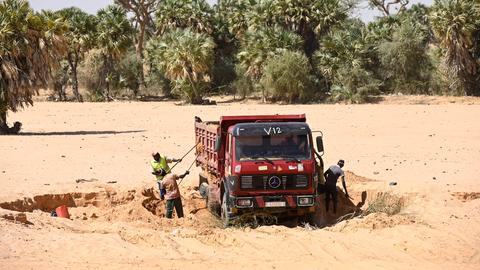Ein Lastwagen auf sandigem Untergrund, Arbeiter schaufeln Sand in das Fahrzeug. Im Hintergrund sind Bäume zu sehen.