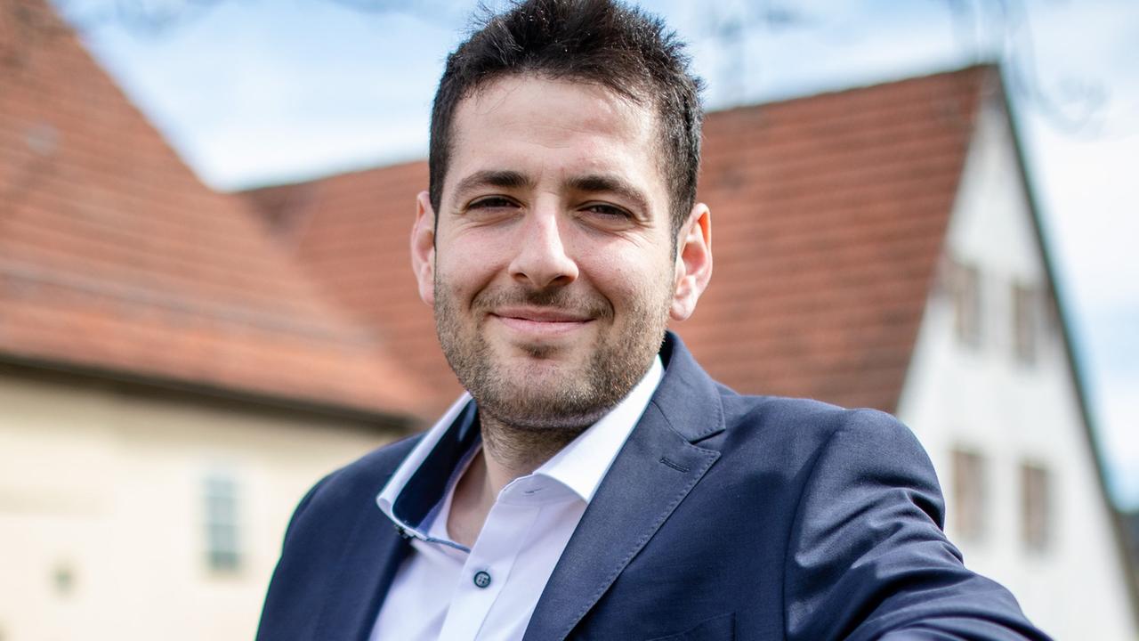 Ein junger, dunkelhaariger Mann, Ryyan Alshebl, sitzt  auf einer Bank und lächelt. 