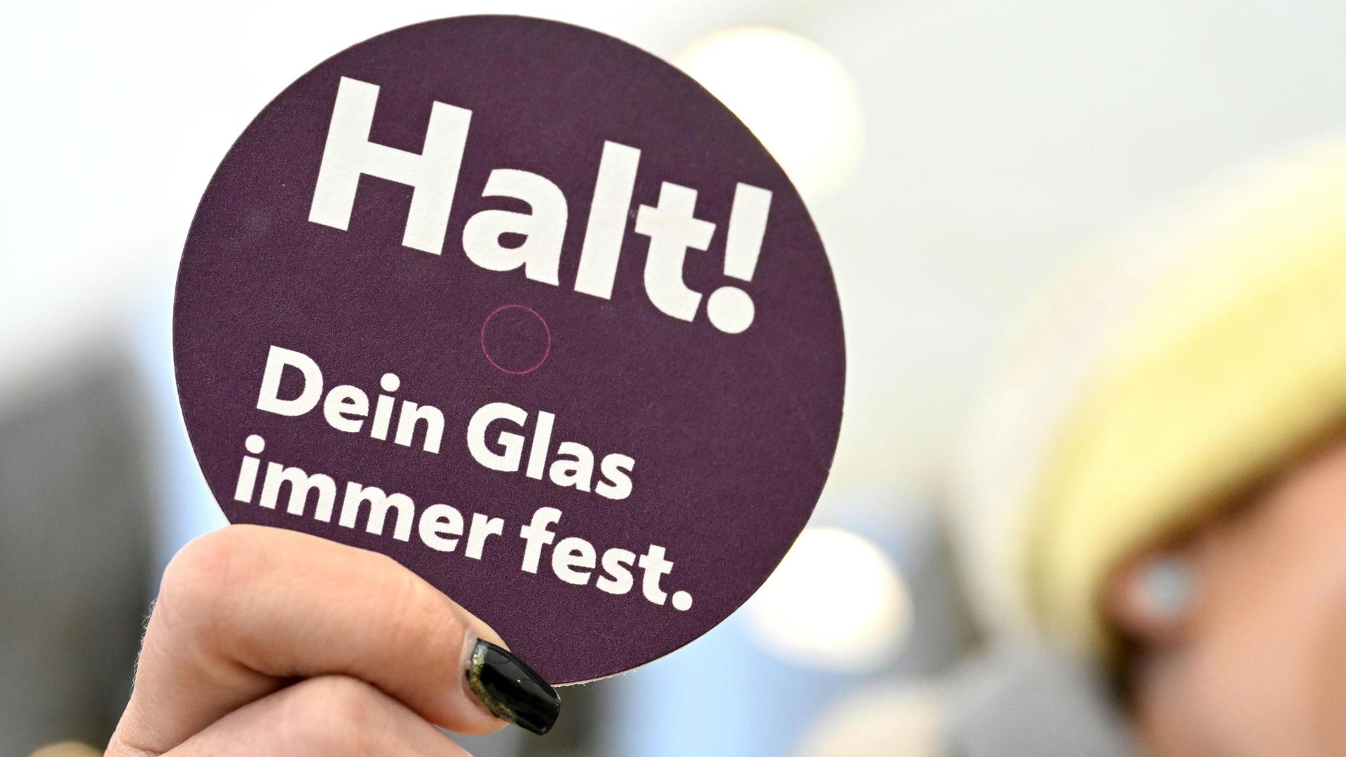 Eine Hand hält ein Schild einer Präventionskampagne zu K.o.-Tropfen hoch auf dem steht: Halt! Dein Glas immer fest.