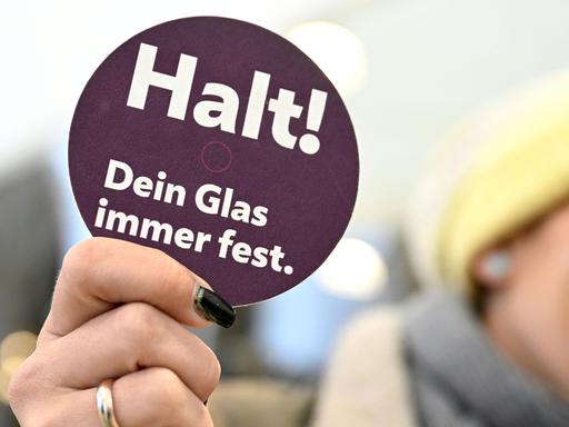 Eine Hand hält ein Schild einer Präventionskampagne zu K.o.-Tropfen hoch auf dem steht: Halt! Dein Glas immer fest.
