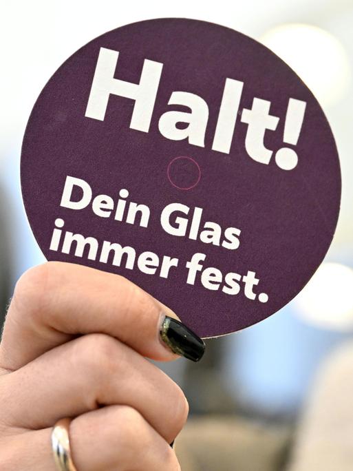 Eine Hand hält ein Schild einer Präventionskampagne zu K.o.-Tropfen hoch auf dem steht: Halt! Dein Glas immer fest.