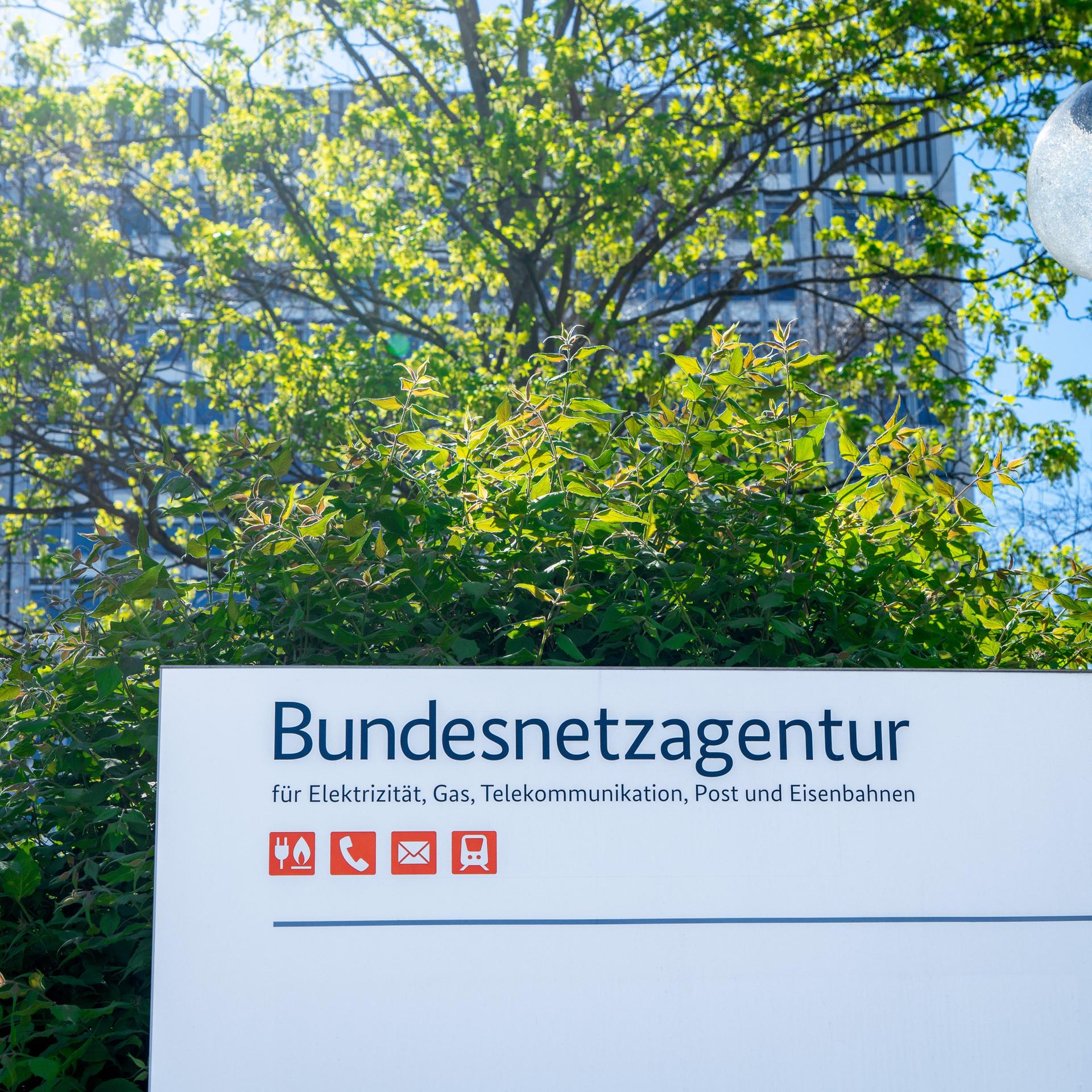 Schild mit der Aufschrift "Bundesnetzagentur", dahinter ein durch einen Baum weitgehend verdecktes Hochhaus.