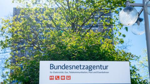 Schild mit der Aufschrift "Bundesnetzagentur", dahinter ein durch einen Baum weitgehend verdecktes Hochhaus.