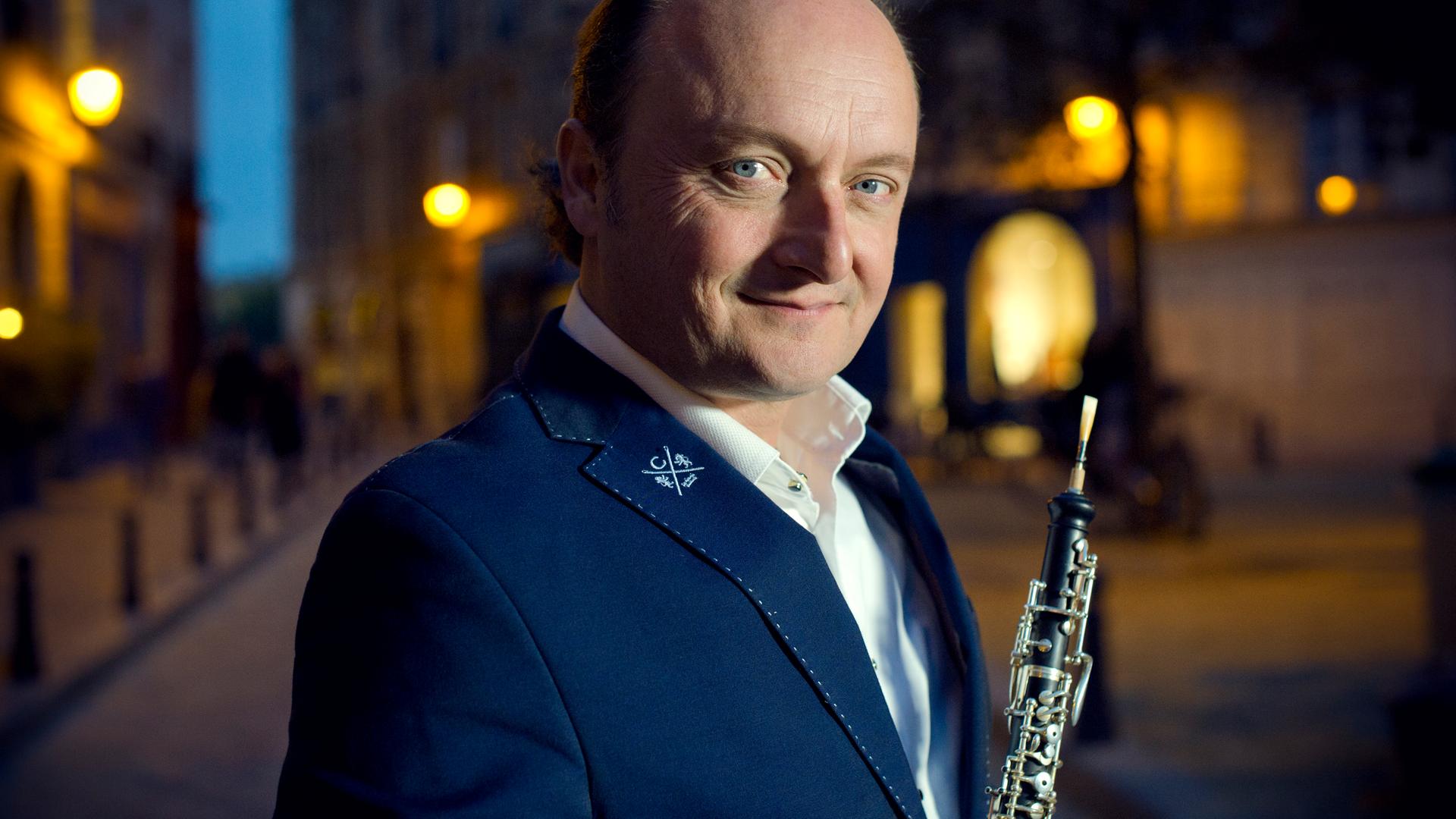 Ein Mann mit Oboe steht in einer nächtlichen Stadt. Er trägt ein weißes Hemd, ein dunkles Sakko und schaut aus blauen Augen freundlich und neugierig in die Kamera. 