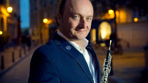 Ein Mann mit Oboe steht in einer nächtlichen Stadt. Er trägt ein weißes Hemd, ein dunkles Sakko und schaut aus blauen Augen freundlich und neugierig in die Kamera. 