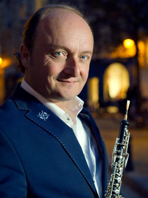Ein Mann mit Oboe steht in einer nächtlichen Stadt. Er trägt ein weißes Hemd, ein dunkles Sakko und schaut aus blauen Augen freundlich und neugierig in die Kamera. 