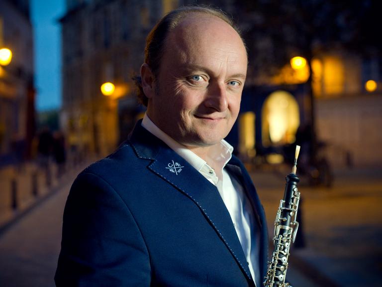 Ein Mann mit Oboe steht in einer nächtlichen Stadt. Er trägt ein weißes Hemd, ein dunkles Sakko und schaut aus blauen Augen freundlich und neugierig in die Kamera. 