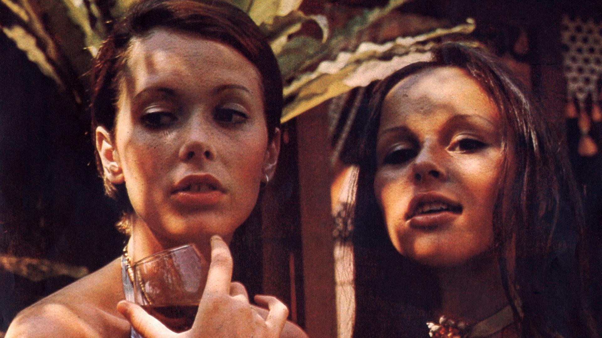 Emanuelle (Sylvia Kristel, links) und Ariane (Jeanne Colletin) aus dem Film "Emanuelle": Sie blicken nach unten, im Hintergrund sind Palmen zu sehen. Die Darstellerin der Emanuelle hält ein halbvolles Rotweinglas in der Hand.  