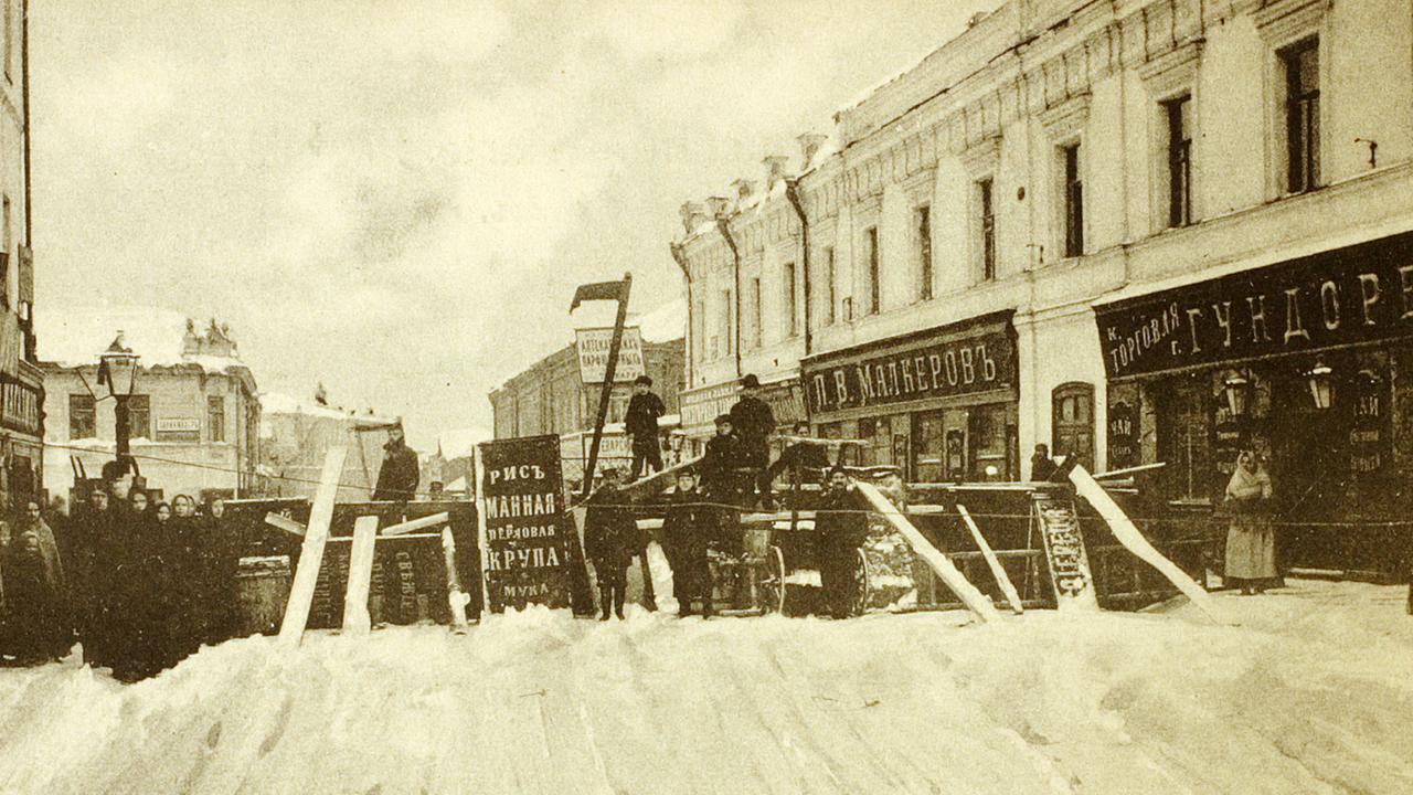 Arbeiteraufstand in Moskau 1905 (Foto). Zu sehenf in schwarz-weiß: Eine Straße mit Barrikaden, an der Seite Häuser mit Geschäften. Arbeiter und Plakate.