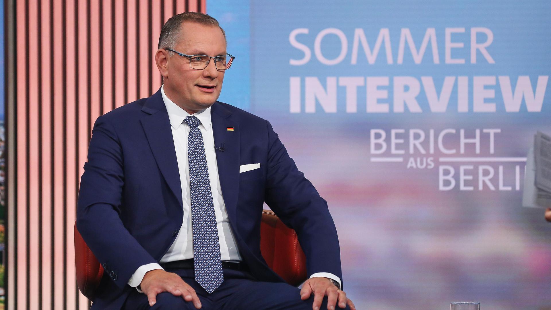 Der AfD-Chef Tino Chrupalla mit dunklem Anzug und Krawatte sitzt auf einem roten Sessel und hält beiden Hände auf den Knien. E sagt gerade etwas. Hinter ihm steht an einer LED-Wand "Sommerinterview - Bericht aus Berlin". 