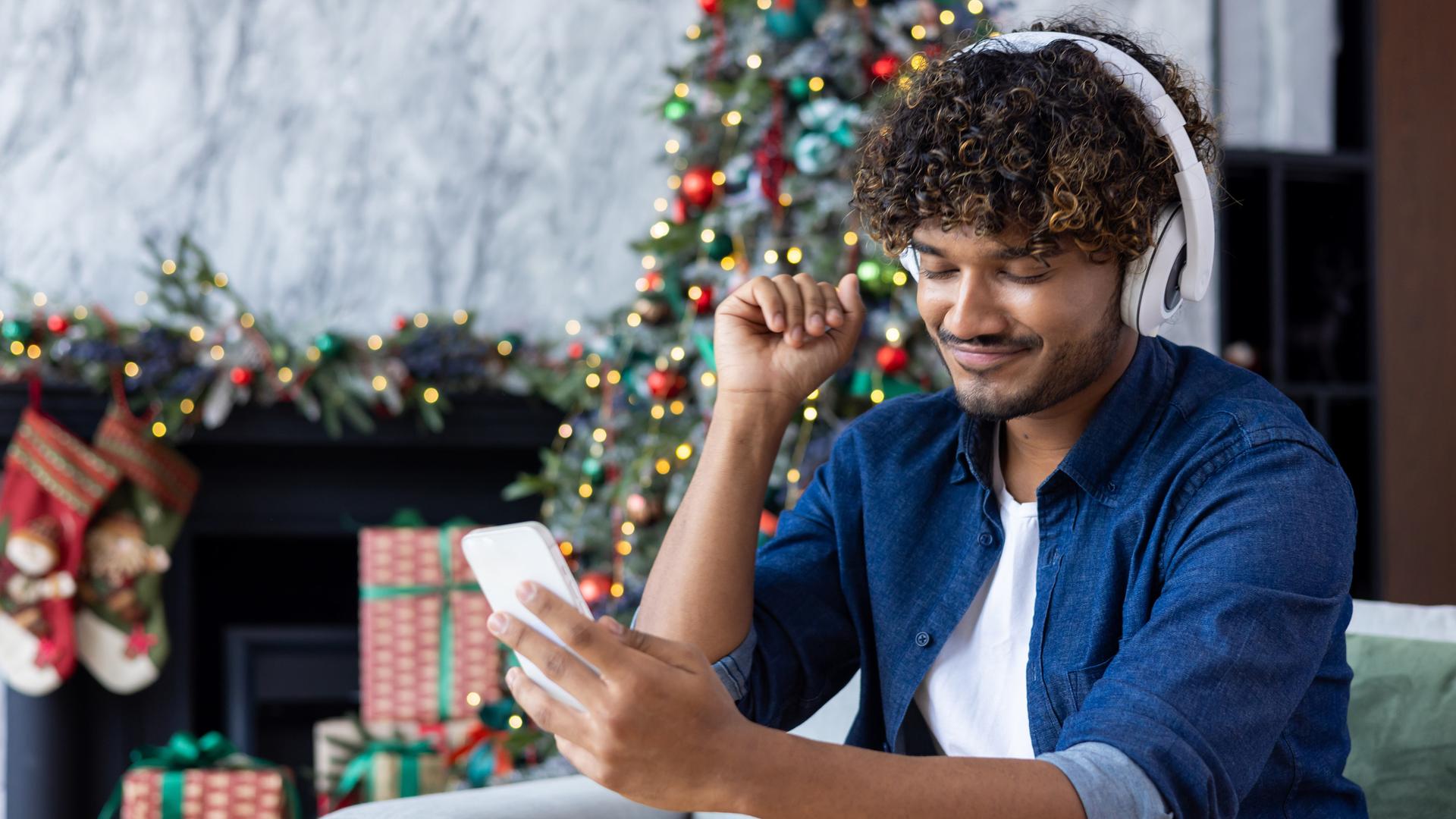 Ein Mann mit Kopfhörern und einem Smartphone in der Hand sitzt in einem Sessel. Im Hintergrund sind ein geschmückter Weihnachtsbaum sowie diverse Geschenke und weitere Dekorationen zu sehen. Ein Mann mit Kopfhörern und einem Smartphone in der Hand sitzt in einem Sessel. Im Hintergrund sind ein geschmückter Weihnachtsbaum sowie diverse Geschenke und weitere Dekorationen zu sehen.