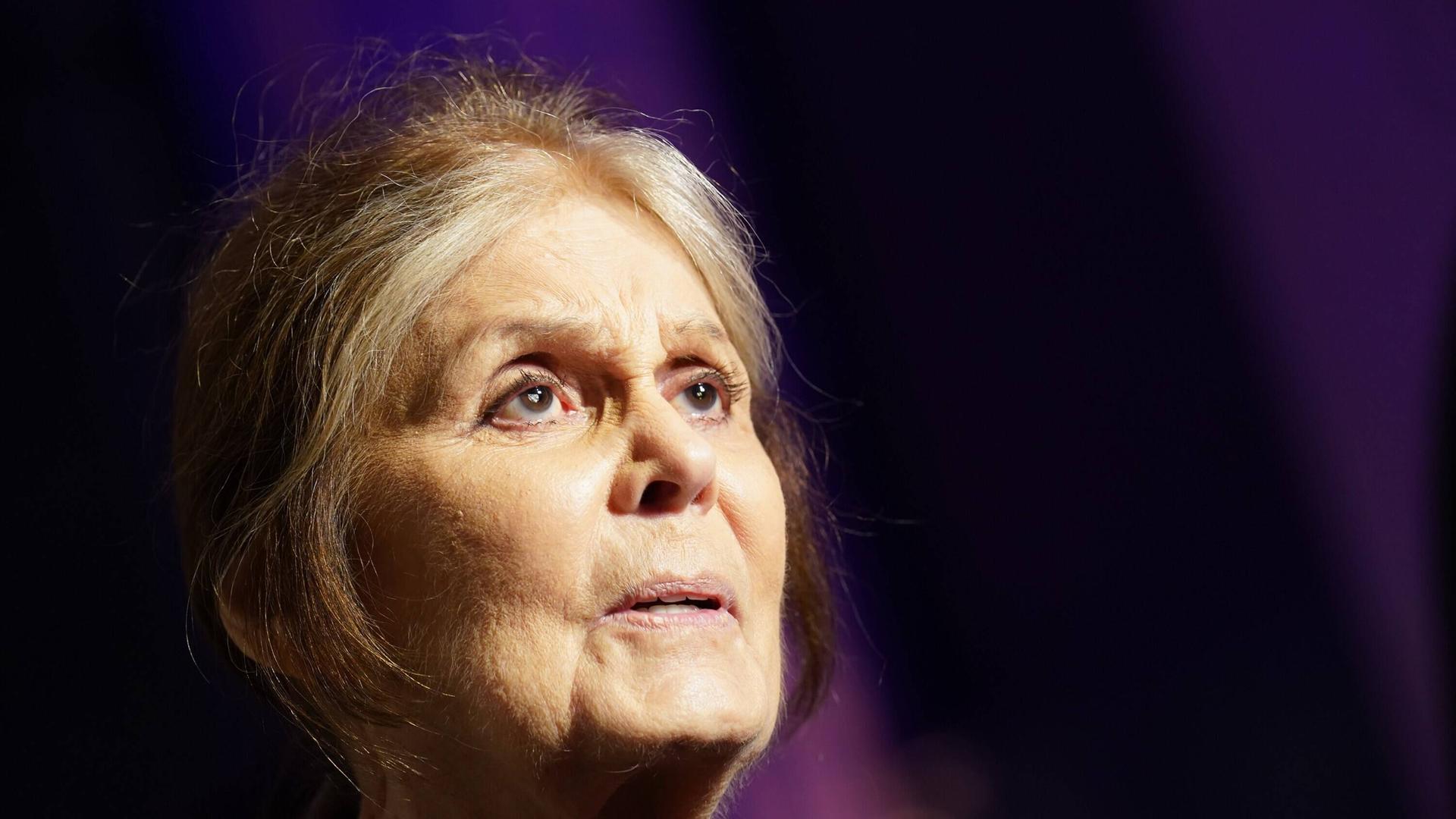 Porträt von Gloria Steinem vor dunklem Hintergrund. Sie schaut mit weit geöffnet Augen nach rechts oben.