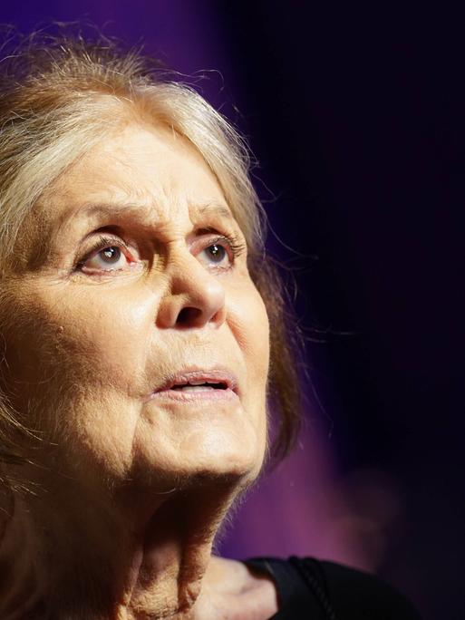 Porträt von Gloria Steinem vor dunklem Hintergrund. Sie schaut mit weit geöffnet Augen nach rechts oben.