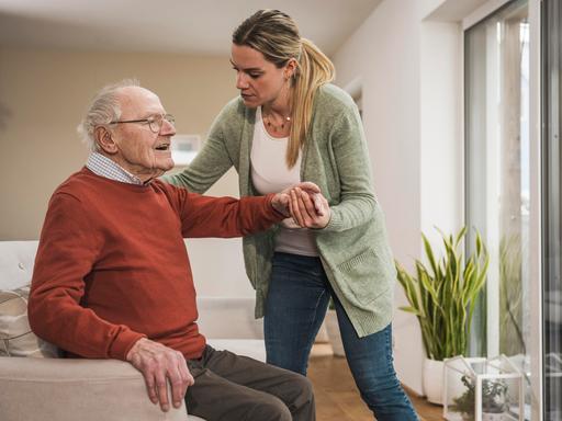 Eine Frau hilf einem Mann in hohem Alter beim Aufstehen aus einem Sessel.