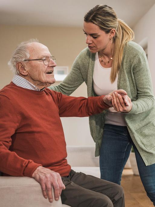 Eine Frau hilf einem Mann in hohem Alter beim Aufstehen aus einem Sessel.