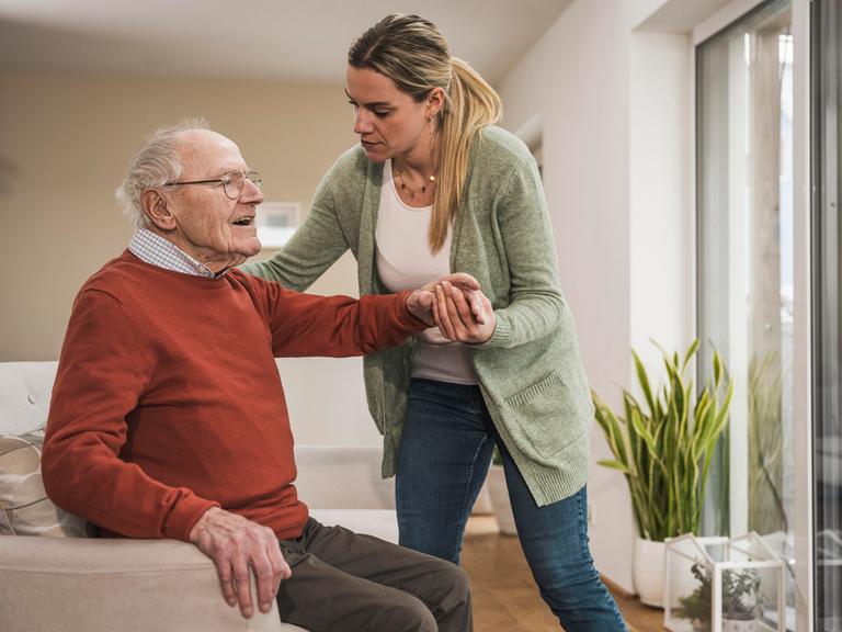 Eine Frau hilf einem Mann in hohem Alter beim Aufstehen aus einem Sessel.