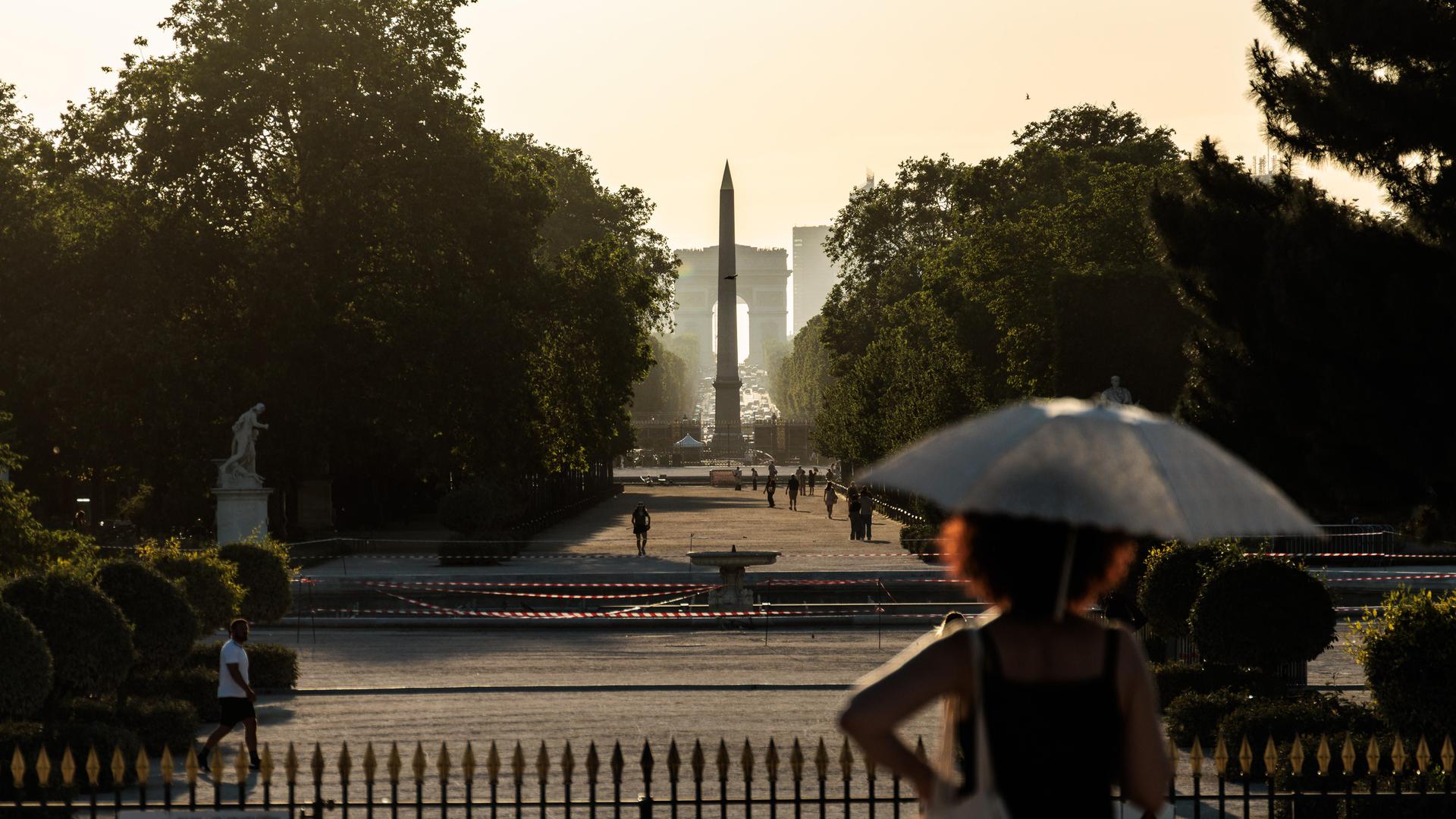 Eine Frau schützt sich während einer intensiven Hitzewelle im Tuileriengarten mit einem Sonnenschirm vor der Sonne – gebadet in goldenes Gegenlicht, mit dem Luxor-Obelisken und dem Arc de Triomphe im Hintergrund – in Paris, Île-de-France, Frankreich.