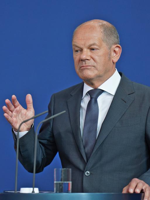 Bundeskanzler Olaf Scholz hebt seine rechte Hand und steht am Pult vor zwei Mikrofonen während einer Pressekonferenz.