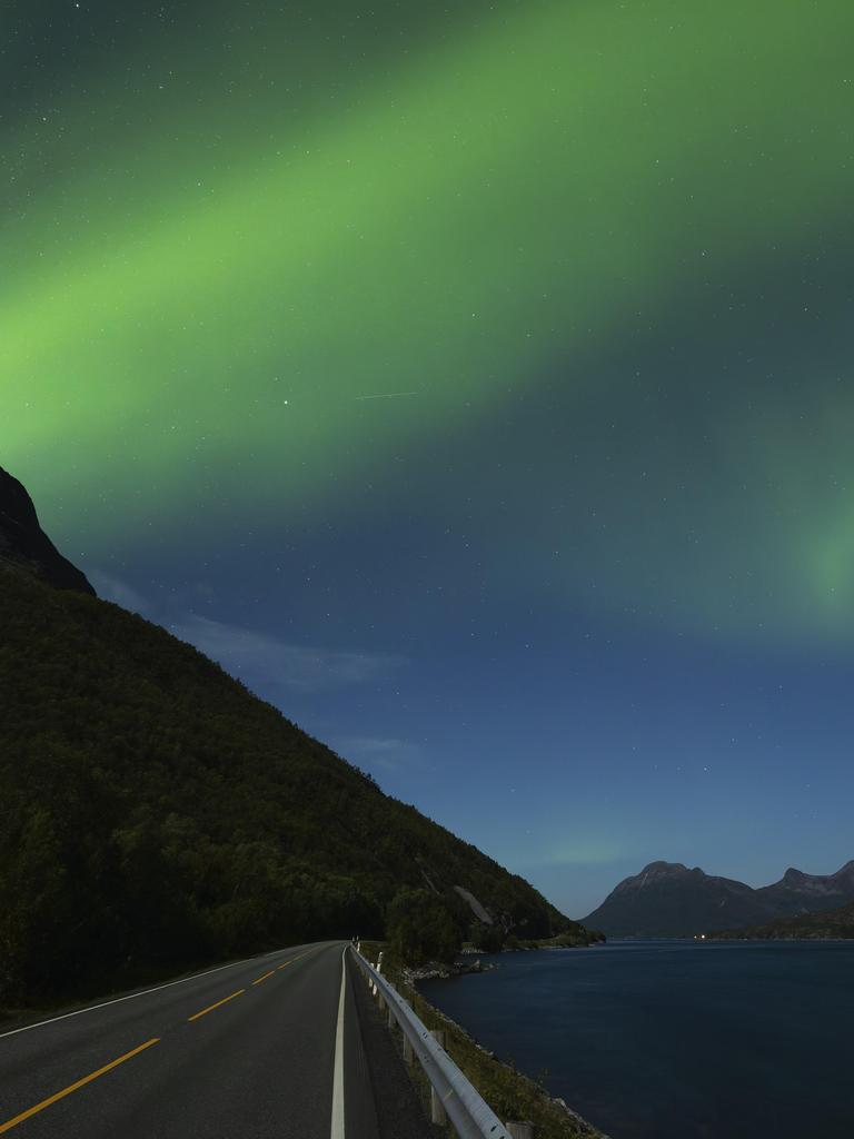 Der Nachthimmel im Norden Norwegens hat sich grün verfärbt. Darunter erstrecken sich Berge und eine Straße, die direkt an einen Fjord grenzt.