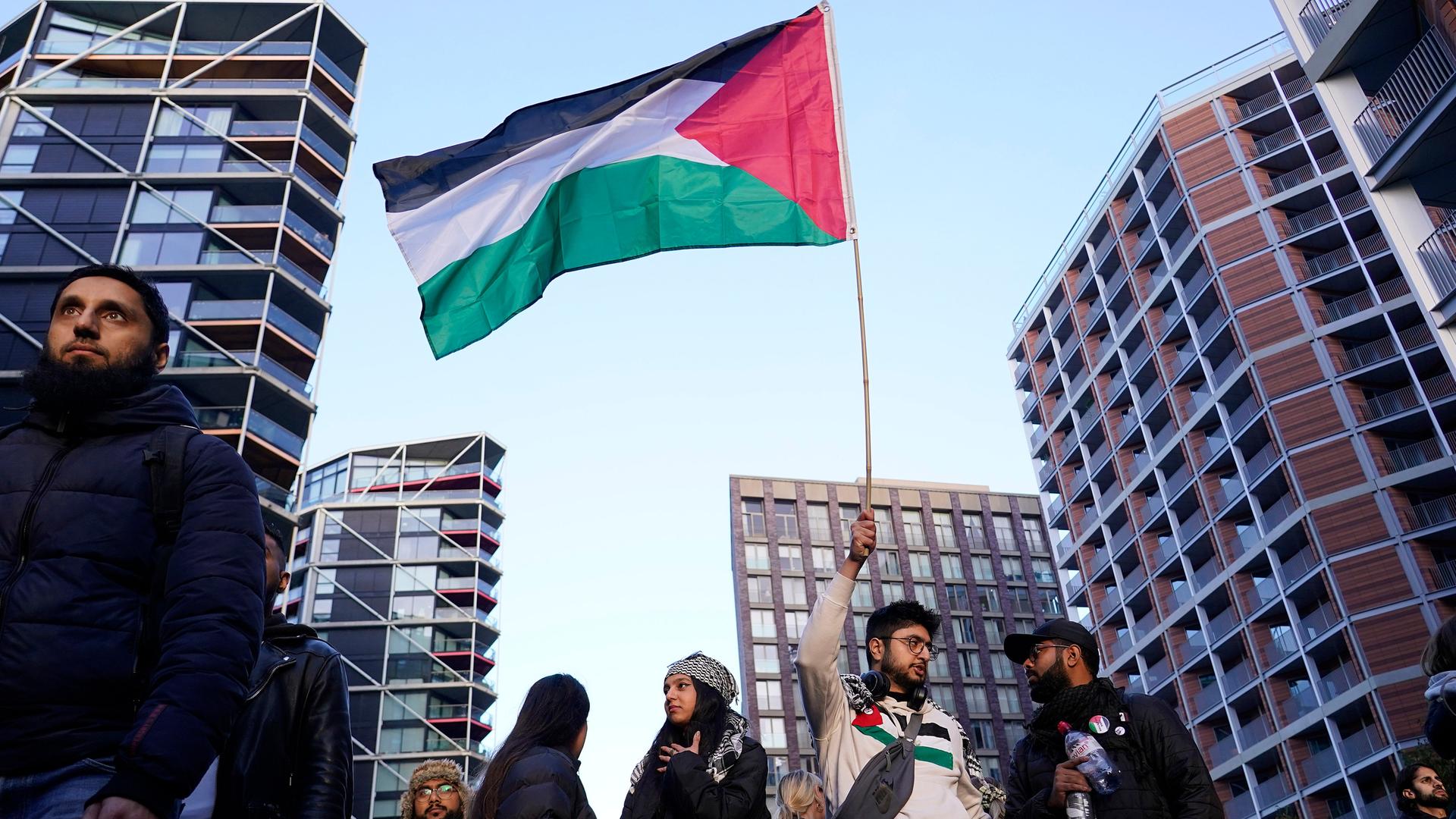 Vor Hochhäusern stehen mehrere Frauen und Männer. Ein Mann hält eine palästinensische Flagge hoch.